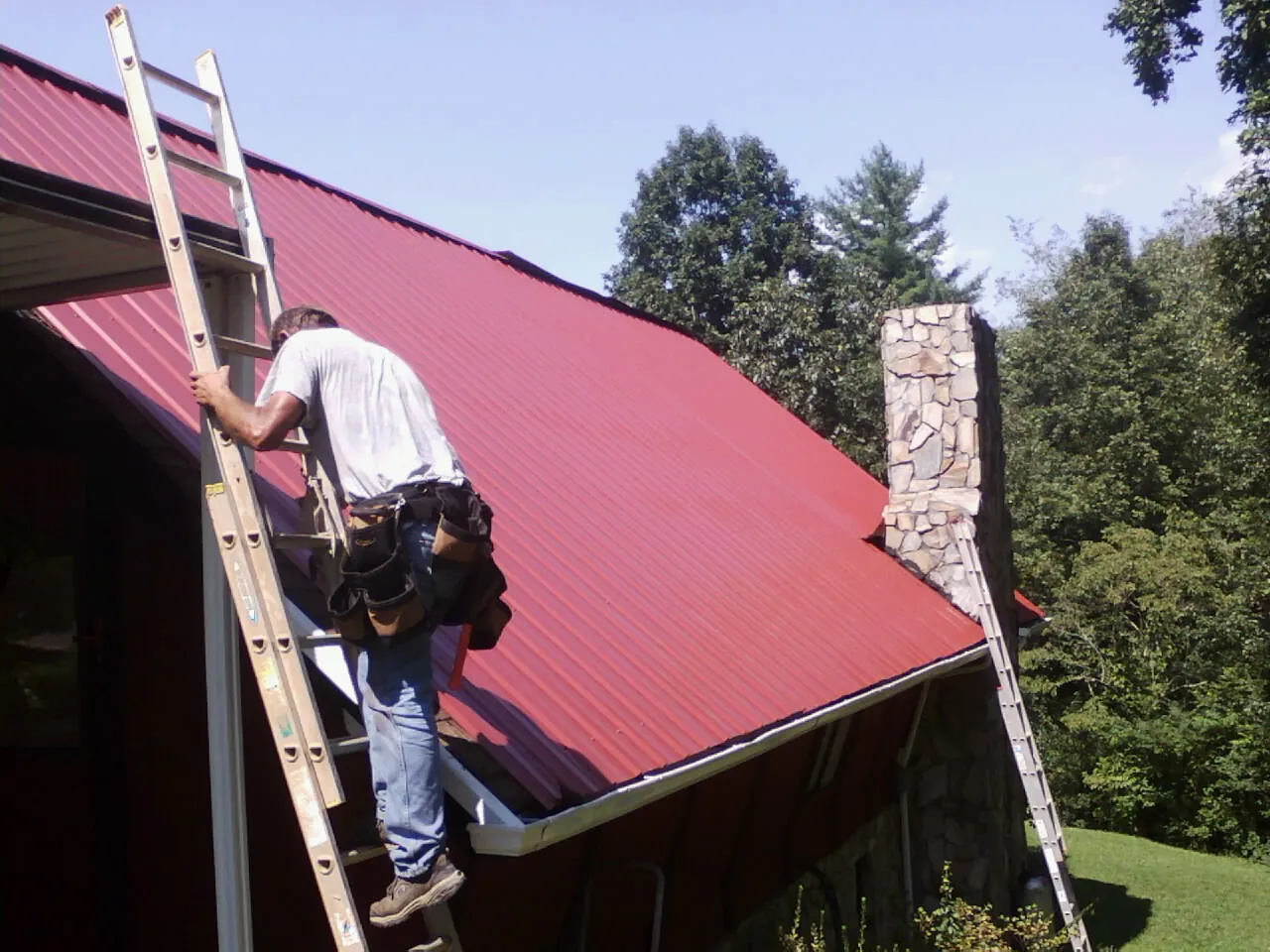 Roofer on a ladder beside a red metal roof and stone chimney — Soffit & Fascia Repair by Roofing Brothers in Belden